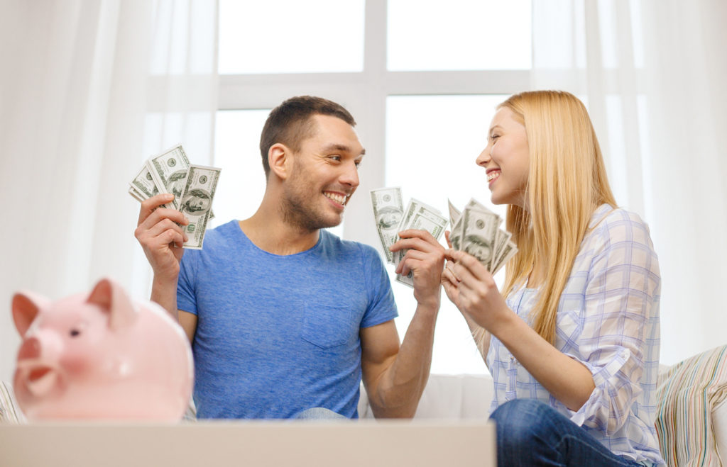 love, family, finance, money and happiness concpet - smiling couple with money and piggybank ot tabl